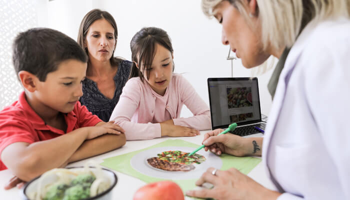 Female dietician at a desk educating a young daughter and son and their mother on healthy eating